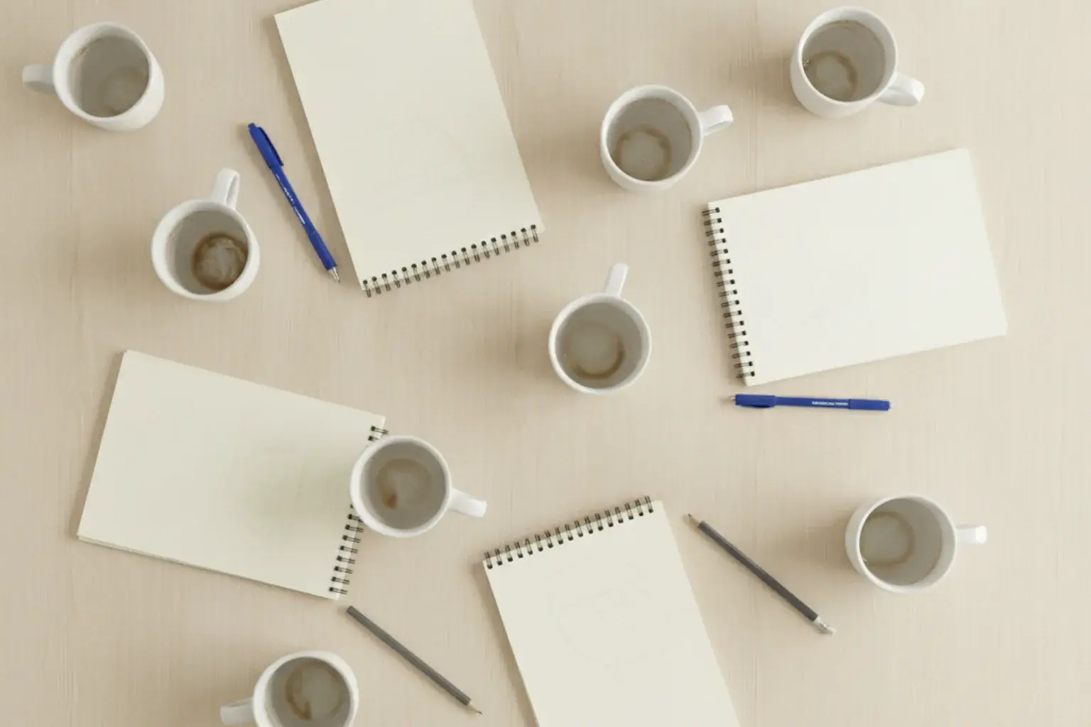 Ein heller Tisch von oben mit mehreren halbvollen Kaffeetassen, leeren Notizblöcken und Stiften, unregelmäßig verteilt